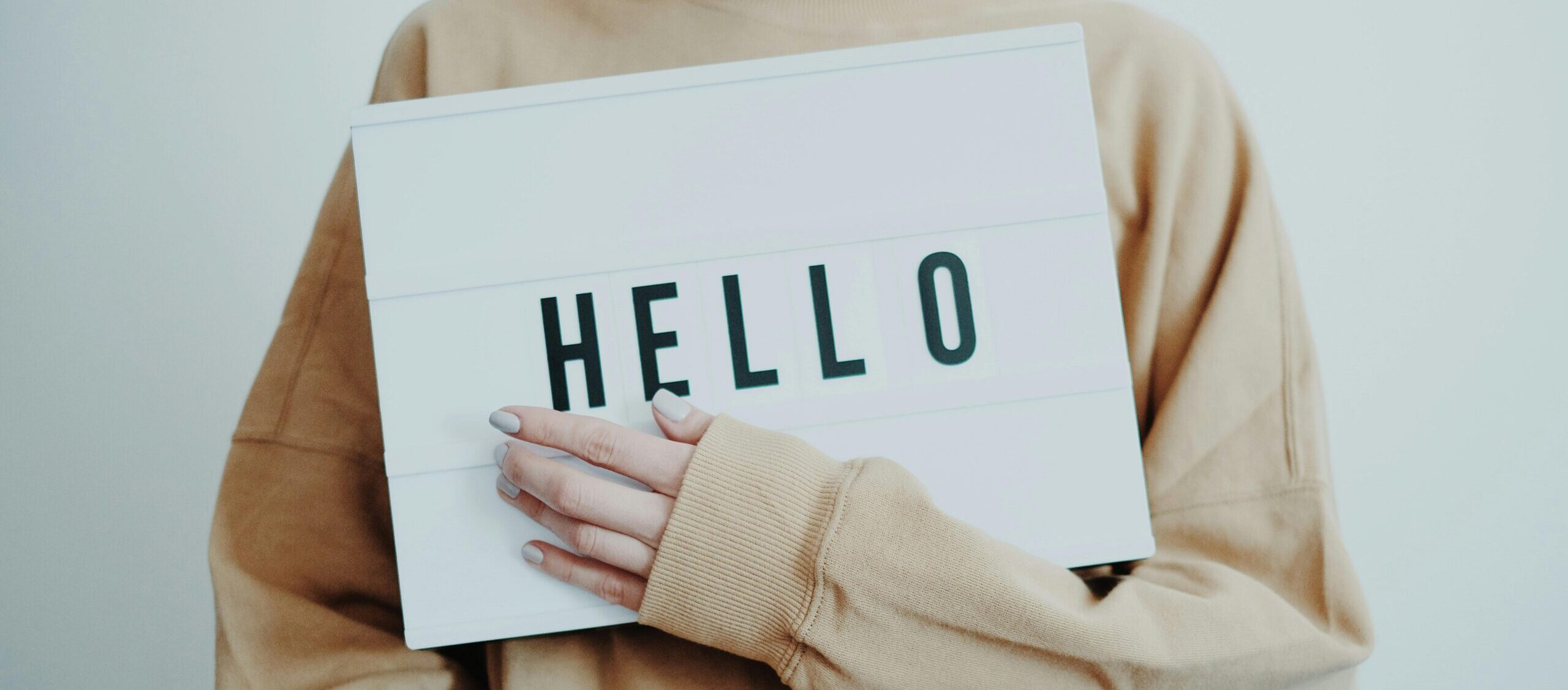 Person in beige sweater holding a hello sign.